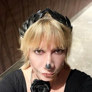 May include: A black feather crown with a silver tiara base. The person is wearing a black top and a black rose necklace. The person has skeleton makeup on their face.