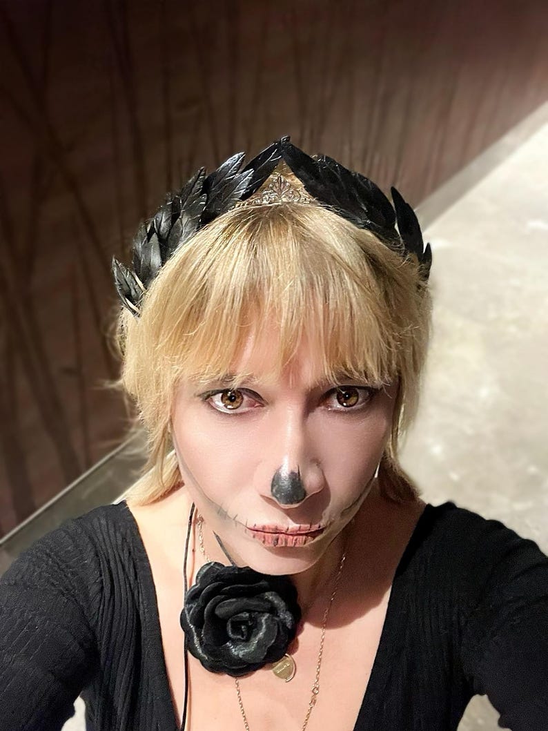 May include: A black feather crown with a silver tiara base. The person is wearing a black top and a black rose necklace. The person has skeleton makeup on their face.