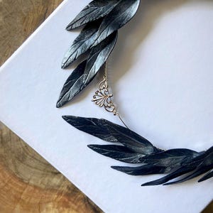 May include: A black leaf-shaped headpiece with a silver chain and decorative pendant. The headpiece is made of multiple dark leaves and is set against a white background. The leaves have a textured appearance.