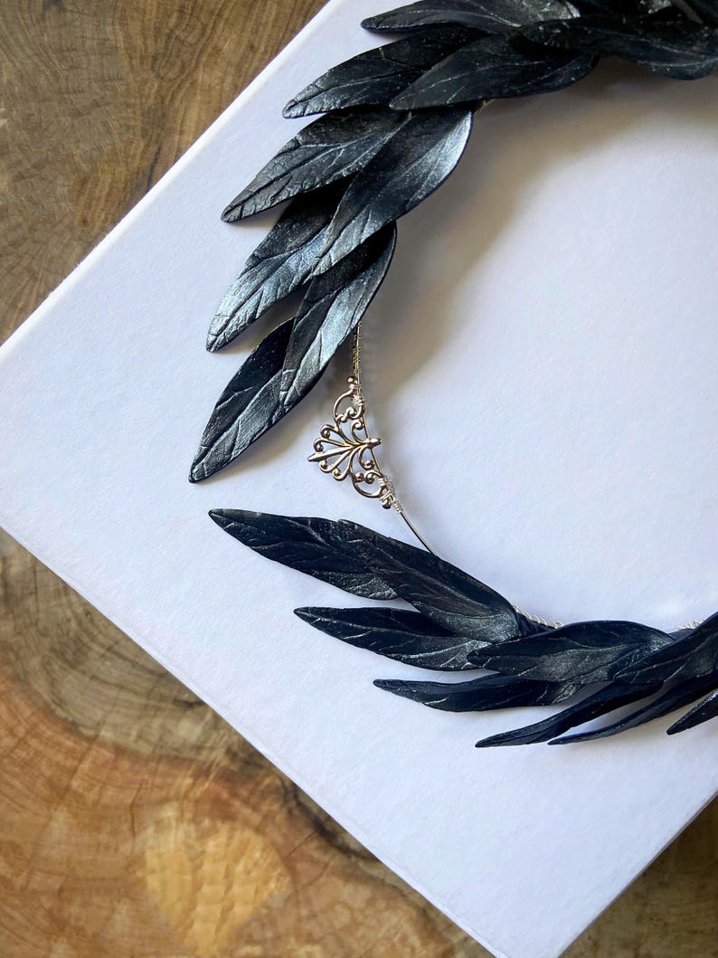 May include: A black leaf-shaped headpiece with a silver chain and decorative pendant. The headpiece is made of multiple dark leaves and is set against a white background. The leaves have a textured appearance.