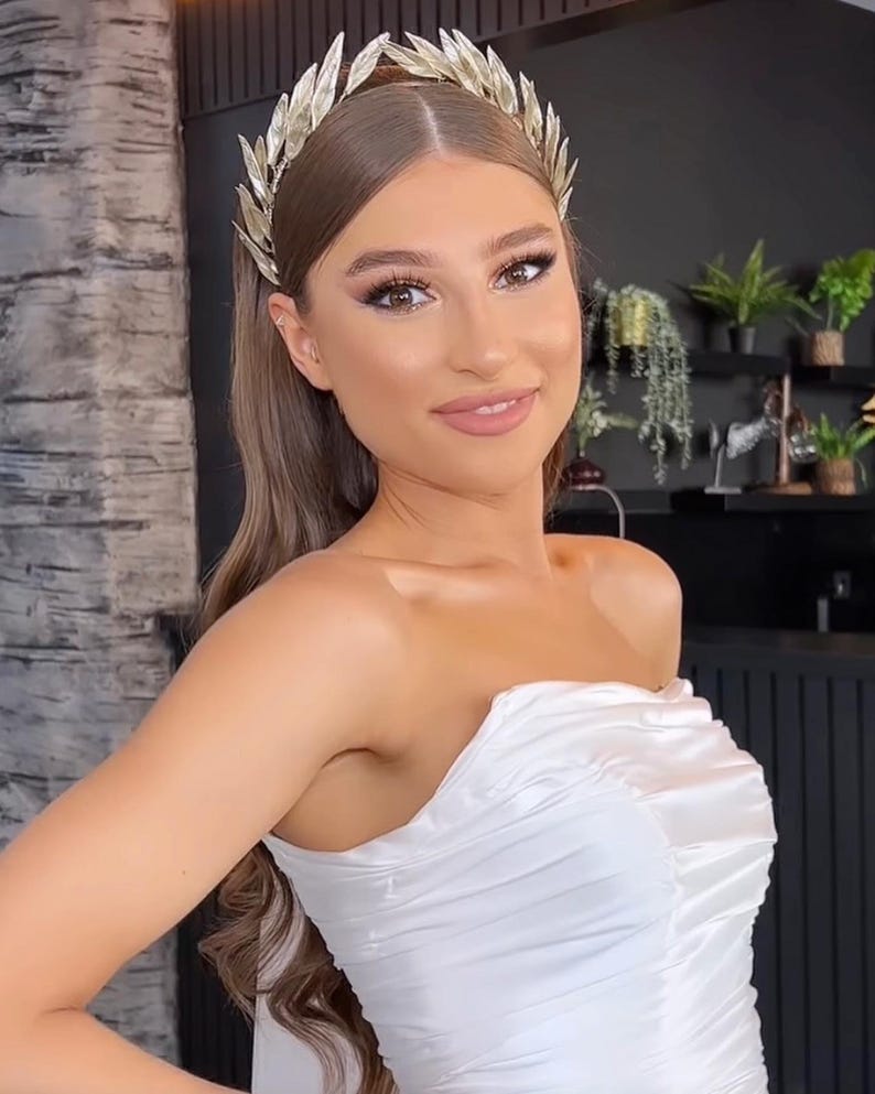 May include: A woman wearing a gold leaf crown and a white strapless dress. The crown is made of delicate, stylized leaves. The dress has a ruched bodice. The background includes a dark wall and plants.