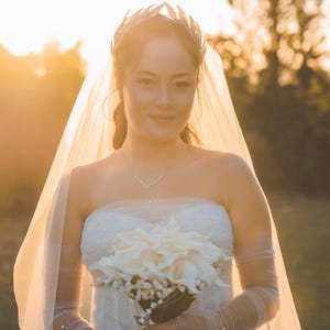 May include: A bride in a strapless white wedding dress, holding a bouquet of white flowers. She wears a silver leaf crown, a delicate necklace, and a sheer veil. The background is a soft, golden sunset.