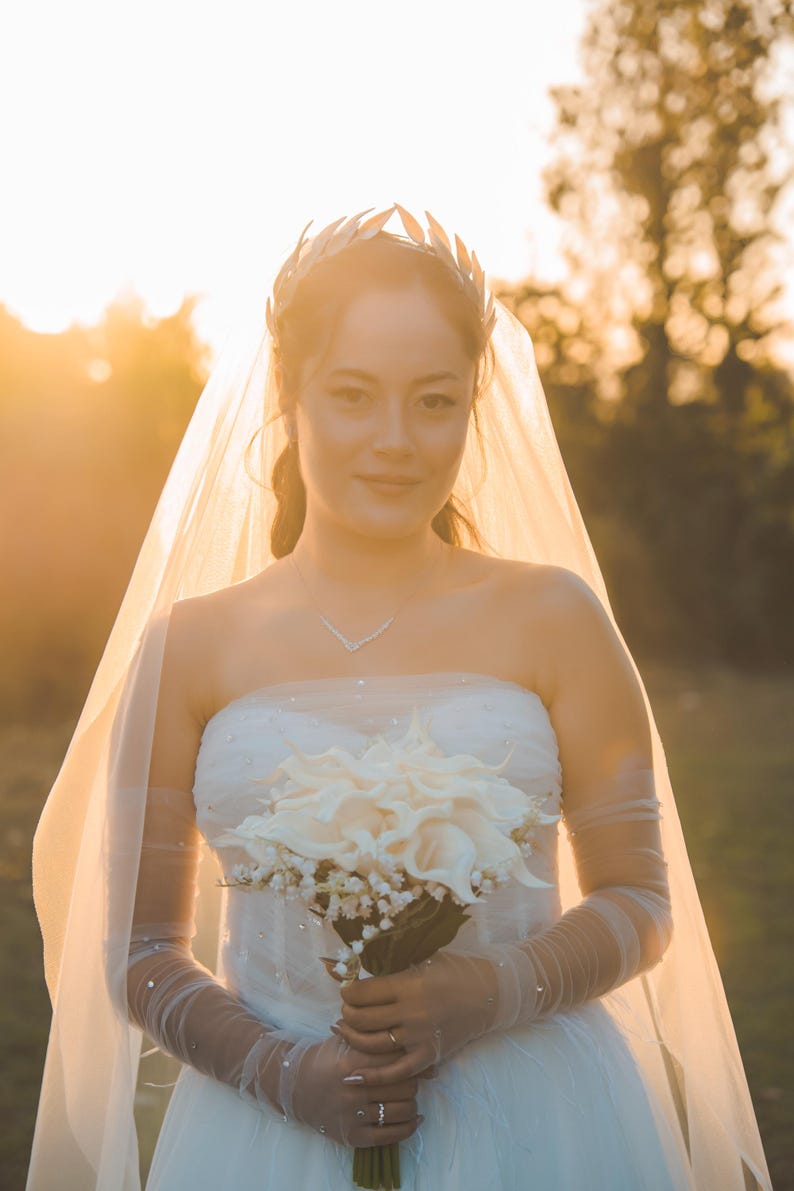 May include: A bride in a strapless white wedding dress, holding a bouquet of white flowers. She wears a silver leaf crown, a delicate necklace, and a sheer veil. The background is a soft, golden sunset.