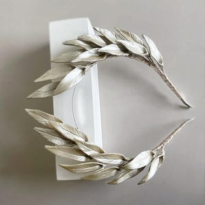 May include: Two silver-colored leaf crowns, possibly for a wedding or special event. The leaves are arranged in a symmetrical pattern, with a textured appearance. The stems are wrapped in a matching silver material. The crowns are displayed against a white block and a neutral background.