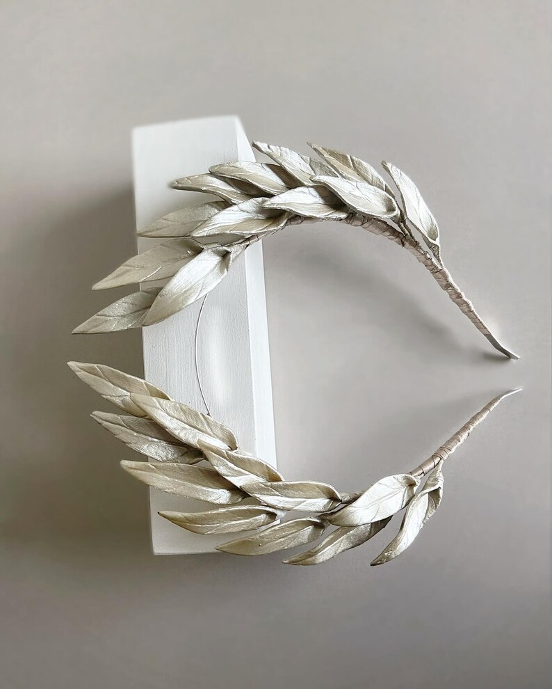 May include: Two silver-colored leaf crowns, possibly for a wedding or special event. The leaves are arranged in a symmetrical pattern, with a textured appearance. The stems are wrapped in a matching silver material. The crowns are displayed against a white block and a neutral background.