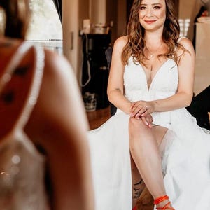 May include: A bride wearing a white wedding dress with a plunging neckline and a flowing skirt. She is wearing a silver crown and orange strappy sandals. The bride is sitting and smiling.