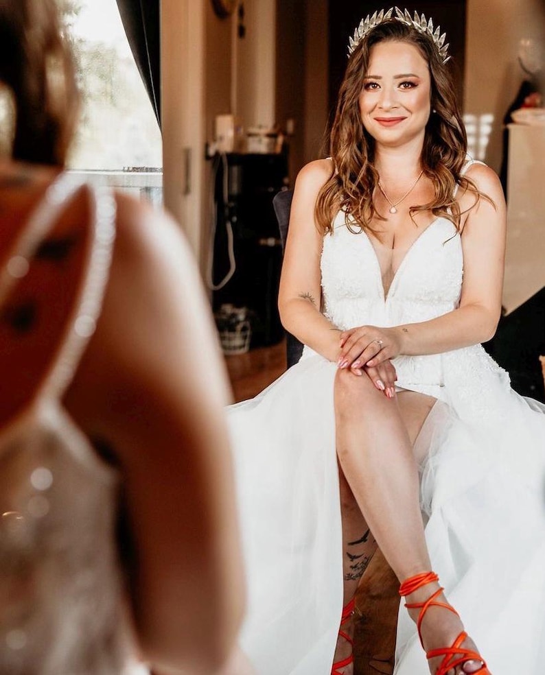 May include: A bride wearing a white wedding dress with a plunging neckline and a flowing skirt. She is wearing a silver crown and orange strappy sandals. The bride is sitting and smiling.