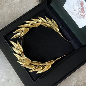 May include: A gold-colored leaf crown, presented in a black box. The crown is made of delicate, detailed leaves, arranged in a circular shape. The box is open, revealing the crown against a black velvet interior. A card with a drawing is visible.