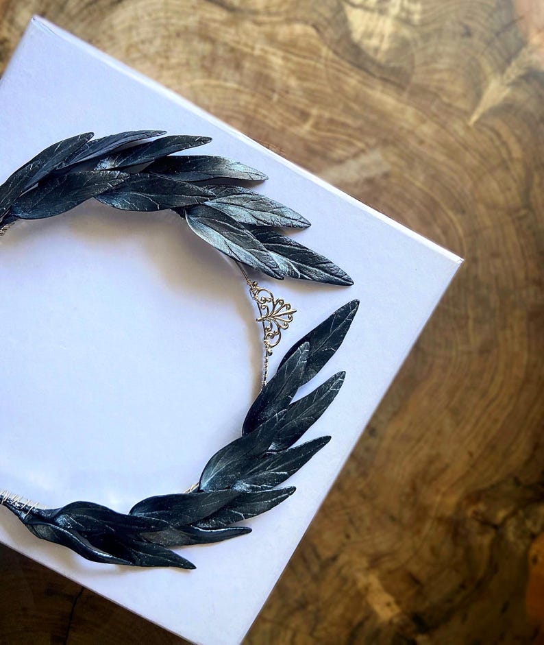 May include: A dark gray leaf crown, likely for a special occasion, rests on a white box. The leaves are detailed and arranged in a circular shape. The background is a wooden surface.