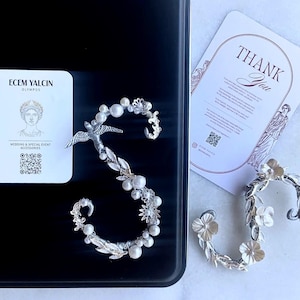 Può includere: Due ear cuff color argento con motivi floreali e di uccelli, impreziositi da perle bianche. Un ear cuff ha la forma della lettera S. Sono incluse due carte bianche con il testo ECEM YALCIN OLYMPOS e THANK YOU, insieme a dei codici QR.