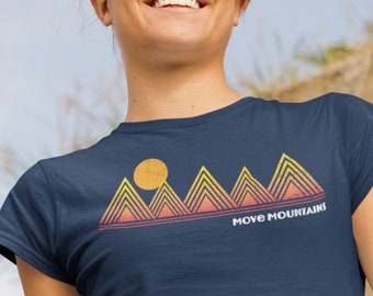 Puede incluir: Una camiseta azul marino con un gráfico de montañas y un sol en estilo retro. El texto "MOVE MOUNTAINS" está impreso debajo del gráfico.