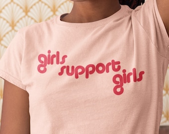 May include: A light pink t-shirt with the text "girls support girls" in a red, bubble-style font. The shirt is worn by a person with dark hair and hoop earrings. The background features a gold and cream-colored geometric pattern.