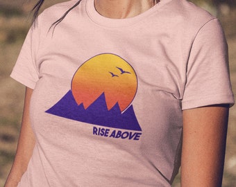 Puede incluir: Una camiseta rosa con un gráfico de una puesta de sol sobre montañas y el texto "RISE ABOVE".