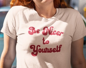 May include: Cream-colored t-shirt with the text "Be Nicer to Yourself" in a retro font. The shirt is short-sleeved and has a crew neck. The text is in shades of red. The person is wearing black jeans and sunglasses.