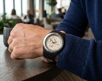May include: A wristwatch with a white face, black band, and silver casing. The watch features red accents on the hands and dials. The band has a textured, perforated design. The watch is on a wrist, with a blue sweater in the background.