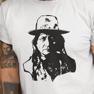 May include: A white t-shirt featuring a black and white illustration of a Native American man wearing a hat. The man has long hair and a beard.