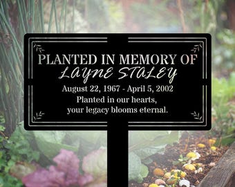 May include: Black metal garden stake with the text "PLANTED IN MEMORY OF Layne Staley" and dates August 22, 1967 - April 5, 2002. The text "Planted in our hearts, your legacy blooms eternal" is also included. The stake is in a garden.