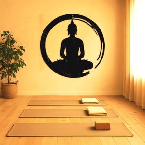 May include: A black silhouette of a meditating Buddha figure inside a circle, on a beige wall. Three brown yoga mats are laid out on a wooden floor.