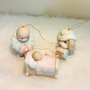 May include: Three porcelain ornaments featuring a child holding a wreath, a child sleeping in a bed with a pink blanket, and a puppy wearing a nightcap holding a mug labeled Cocoa 1998. Each ornament has a gold hanging string attached.