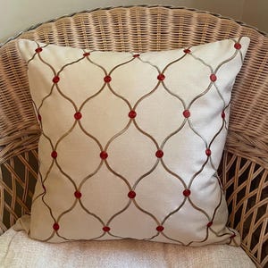 May include: A square decorative pillow with a cream-colored background and an embroidered pattern of intersecting lines and red circles. The pillow is placed on a wicker chair.