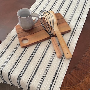 May include: A white and black striped table runner with a wooden cutting board, a ceramic mug, and a wooden whisk and spatula.