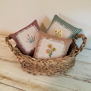 May include: A woven basket filled with three small, square pillows. The pillows have a gingham pattern in burgundy, brown, and green. Each pillow features a linen patch with embroidered floral designs in yellow, pink, and green.