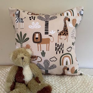 May include: A beige square pillow with a safari animal print featuring a zebra, lion, and giraffe. A brown teddy bear sits in front of the pillow. The pillow has a soft, textured appearance, and the bear is made of plush material.