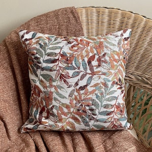 May include: A white throw pillow with a colorful floral pattern featuring shades of orange, red, and blue. The pillow is sitting on a wicker chair with a brown throw blanket.