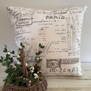 May include: A decorative pillow with a vintage Parisian design. The off-white pillow features faded black script, stamps, and text, including "PARIS" and "PONTOISE." A woven basket with greenery sits in front of the pillow.