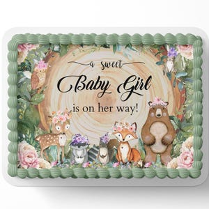 May include: A green and white cake with a woodland theme. The cake is decorated with a brown bear, a deer, a fox, and a squirrel. The cake is topped with a message that says "A sweet baby girl is on her way!"