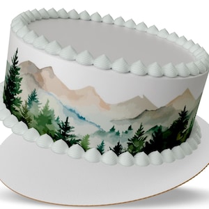 May include: A white cake with a mountain and forest design. The cake features a watercolor landscape with green trees and tan mountains. White frosting lines the top and bottom edges of the cake.
