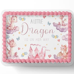 May include: A rectangular cake with pink frosting trim and a white center. The center features a watercolor design with pink dragons, a pink castle, clouds, hearts, stars, and the text "A Little Dragon Is On Her Way."