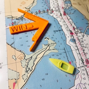 May include: An orange plastic wind direction tool and a yellow boat-shaped tool rest on a nautical chart. The wind tool has the word "WIND" printed on it. The chart shows coastal features, depths, and navigational markers.