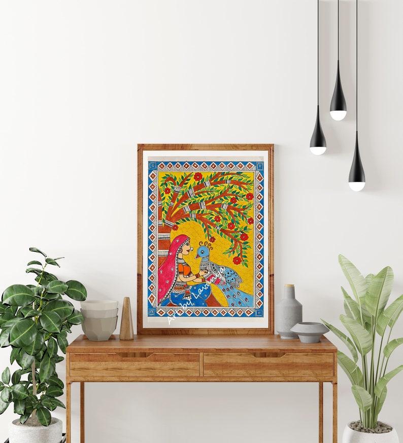 May include: A framed painting of a woman in a colorful sari sitting under a tree with a peacock. The painting is done in a folk art style with bright colors and simple lines.
