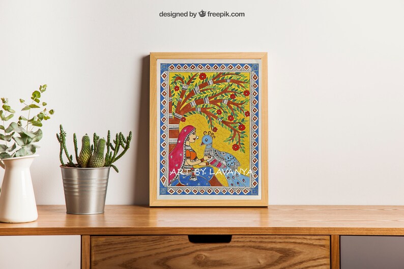 May include: A framed painting of a woman in a red and blue dress sitting under a tree with a peacock. The painting is in a traditional Indian style with bright colors and intricate details. The text "ART BY LAVANYA" is at the bottom of the painting.