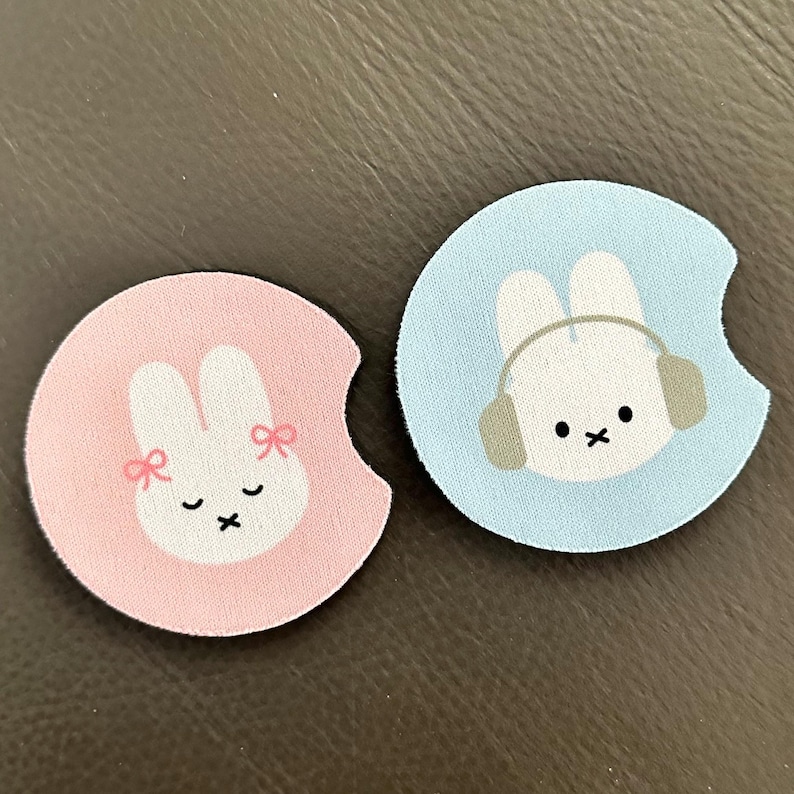 May include: Two round pink and blue coasters with a white bunny design. The pink coaster has a bunny with pink bows and the blue coaster has a bunny wearing headphones.