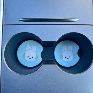 May include: Two blue cup holders with a white bunny wearing headphones design. The cup holders are in a car.