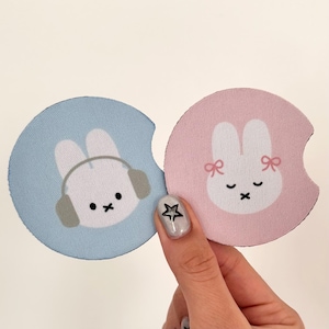 May include: Two round fabric coasters with bunny designs. One coaster is light blue with a white bunny wearing headphones. The other coaster is pink with a white bunny with pink bows on its ears.