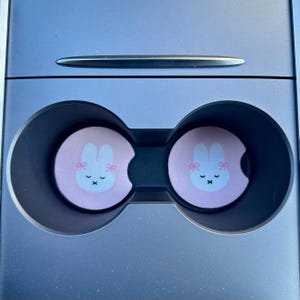 May include: Two pink car cup holders with a white bunny with pink bows and a pink nose. The bunny has a closed mouth and a small X for eyes.