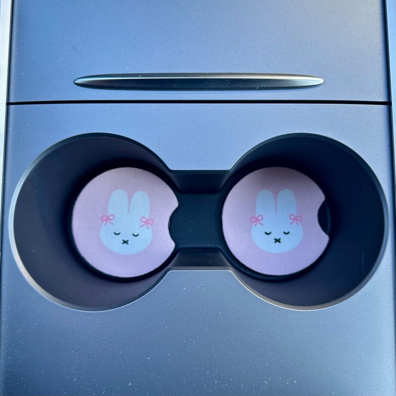 May include: Two pink car cup holders with a white bunny with pink bows and a pink nose. The bunny has a closed mouth and a small X for eyes.