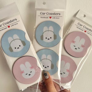 May include: Set of six car coasters featuring a cute bunny design. Two coasters are light blue with a bunny wearing headphones, two are pink with a bunny wearing a bow, and two are light blue with a bunny wearing a headband. The coasters are handmade with love.