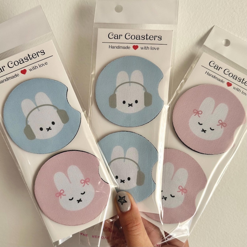 May include: Set of six car coasters featuring a cute bunny design. Two coasters are light blue with a bunny wearing headphones, two are pink with a bunny wearing a bow, and two are light blue with a bunny wearing a headband. The coasters are handmade with love.