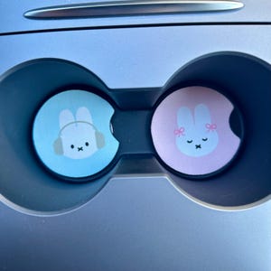 May include: Two car cup holders with a blue and pink bunny design. The blue bunny is wearing headphones and the pink bunny has pink bows.