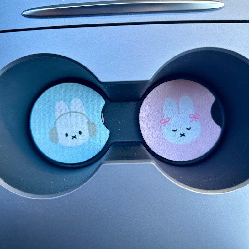 May include: Two car cup holders with a blue and pink bunny design. The blue bunny is wearing headphones and the pink bunny has pink bows.