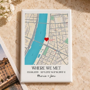 May include: A rectangular white plaque featuring a printed map with a red heart icon marking a specific location. Below the map, the text reads WHERE WE MET, 03.08.2019, 50°0.3112' N 8°16.3913' E, and the names Marie + Joni.