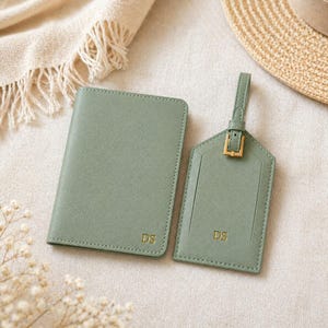 May include: Sage green passport holder and luggage tag set. The passport holder and tag are made of a textured material and feature gold-colored initials "DS". The luggage tag has a buckle closure and a strap. The set is displayed on a neutral background.