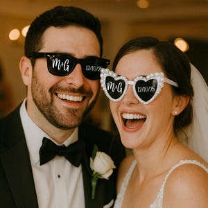Può includere: Una coppia sorridente indossa occhiali da sole originali. L'uomo indossa occhiali da sole neri con "M+G" e "12/10/25" stampati. La donna indossa occhiali da sole a forma di cuore con perle e lo stesso testo.