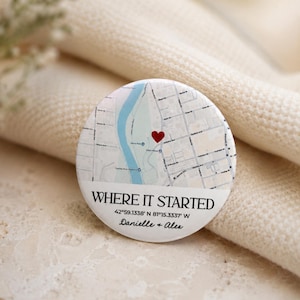 Può includere: Un bottone rotondo bianco con un design a mappa e un cuore rosso che segna una posizione. Il testo "WHERE IT STARTED" è sopra le coordinate 42°59.1338' N 81°15.3337' W, con i nomi "Danielle + Alex" sotto. Il bottone è su una superficie strutturata.