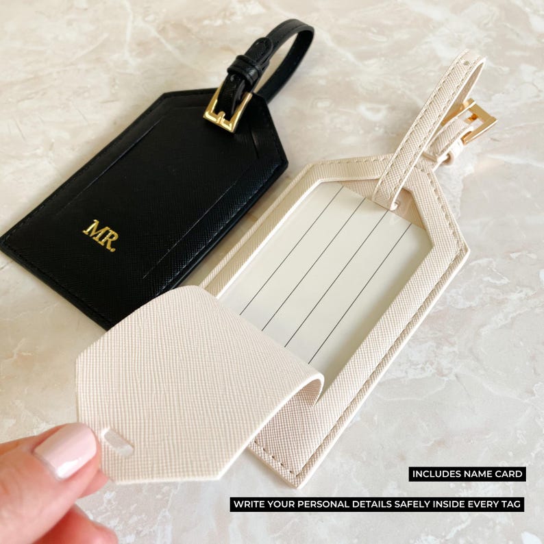 Peut inclure: Deux &eacute;tiquettes de bagage, une noire et une cr&egrave;me, avec des accessoires dor&eacute;s. L'&eacute;tiquette noire porte l'inscription "MR." en lettres dor&eacute;es. L'&eacute;tiquette cr&egrave;me a une fen&ecirc;tre transparente pour une carte de nom. Le texte sur l'image indique : "INCLUDES NAME CARD" et "WRITE YOUR PERSONAL DETAILS SAFELY INSIDE EVERY TAG."