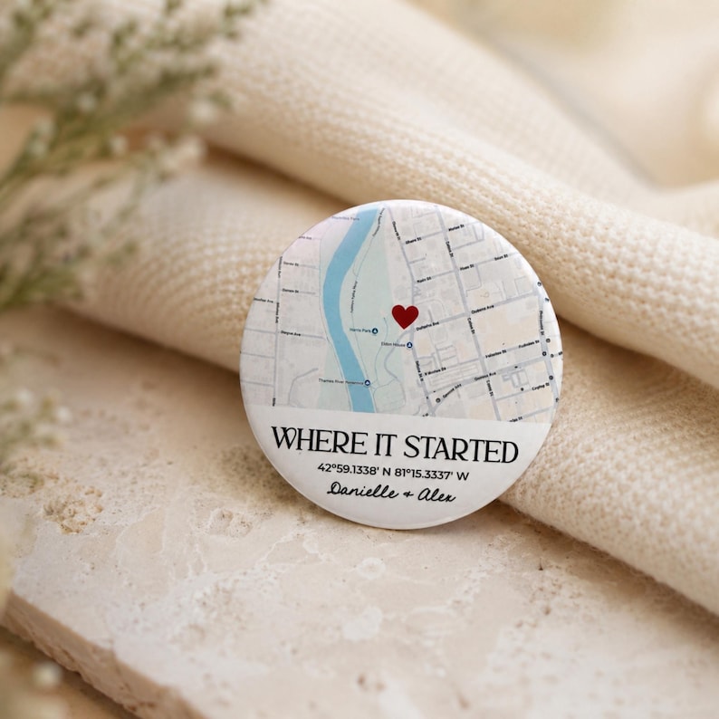 May include: A round, white button with a map design and a red heart marking a location. The text "WHERE IT STARTED" is above the coordinates 42&deg;59.1338' N 81&deg;15.3337' W, with the names "Danielle + Alex" below.